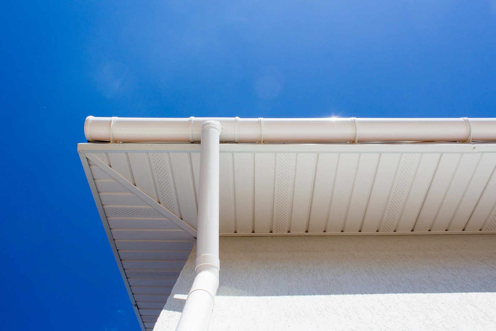 White gutter and downspout along a finished white soffit and wall under a blue sky.