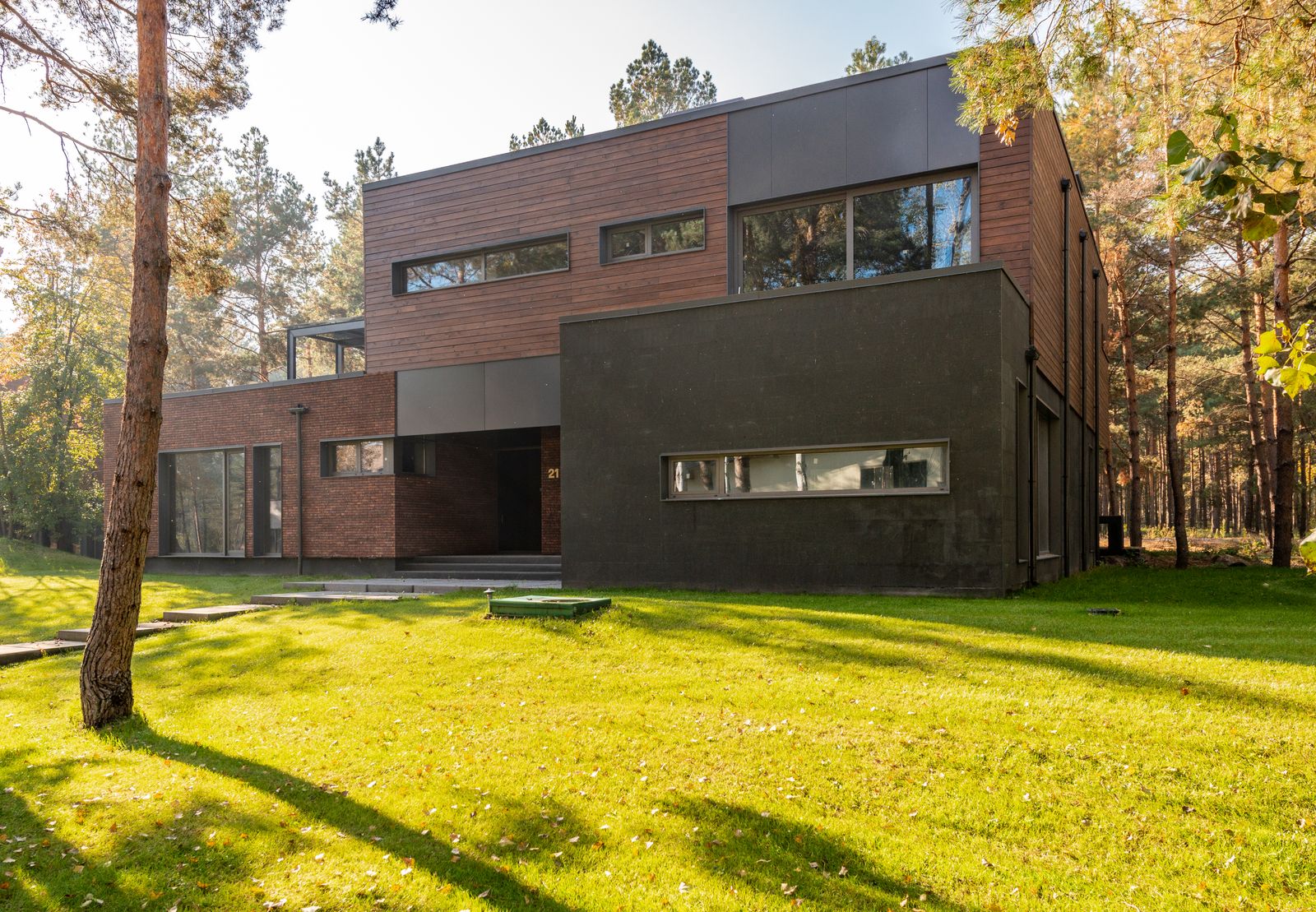 Finished contemporary home among trees, with wide dark-framed windows, wood-tone upper cladding and a dark lower facade.
